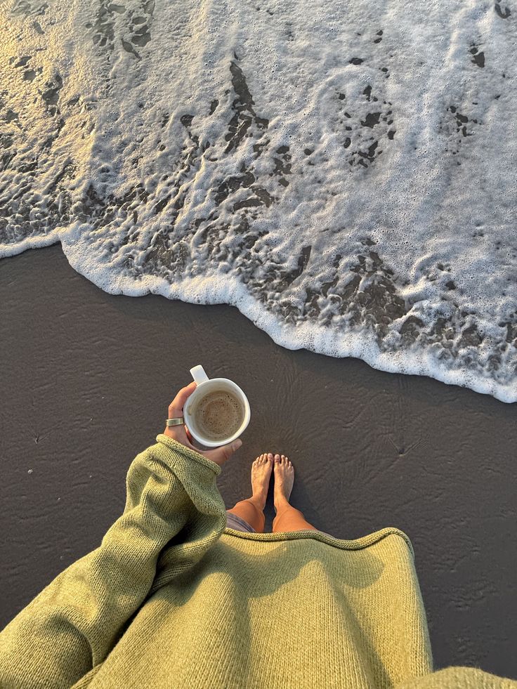 Foto primeira pessoa na praia: uma mão segurando uma caneca de café diante da espuma do mar